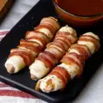 bacon wrapped in cheese on a black plate with dipping sauce next to the bowl full of ketchup