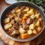a pot filled with stew and carrots on top of a white countertop next to a wooden spoon