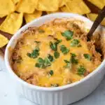 a hand holding a tortilla chip over a bowl of chili and cheese dip