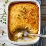 a white casserole dish with cheese and potatoes in it, next to a wooden spoon